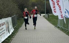 Zdjęcie przedstawia moment, w kt&oacute;rym dw&oacute;ch biegaczy przekracza linię mety zawod&oacute;w biegowych. Trasa wyłożona jest kostką brukową, a po bokach znajduje się metalowe ogrodzenie oraz banery promocyjne. Na g&oacute;rze kadru widoczny jest baner z logotypem &bdquo;Śremski S
