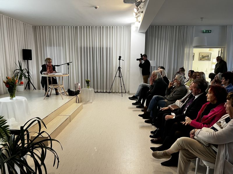 Na fotografii wykonanej w jasnym, nowocześnie urządzonym wnętrzu widać wydarzenie kulturalne, które odbywa się w sali Biblioteki Publicznej im. Heliodora Święcickiego w Śremie. Po lewej stronie zdjęcia, na niewielkim podwyższeniu, przy okrągłym stole zasiada dr Danuta Płygawko. Ubrana jest w ciemną marynarkę i różowy szalik, a przed sobą ma mikrofon i notatki. W tle za nią rozciąga się biała zasłona zakrywająca całą ścianę. Na prawo od sceny widzimy publiczność – kilkanaście osób siedzących na krzesłach w kilku rzędach. Większość słuchaczy to osoby starsze. Słuchają z uwagą, część ma założone ręce, inni trzymają programy lub książki. W środku sali stoi statyw z kamerą skierowaną na prowadzącą spotkanie. Obok niej, przy ścianie, mężczyzna z aparatem fotograficznym robi zdjęcia. Cała scena jest dobrze oświetlona, wnętrze urządzone minimalistycznie – jasne ściany, białe zasłony, subtelne oświetlenie sufitowe. Przed sceną stoją dwa okrągłe stoliki przykryte białymi obrusami, na których znajdują się wazony z kolorowymi tulipanami – wprowadzają one akcent wiosennego ciepła do wnętrza.