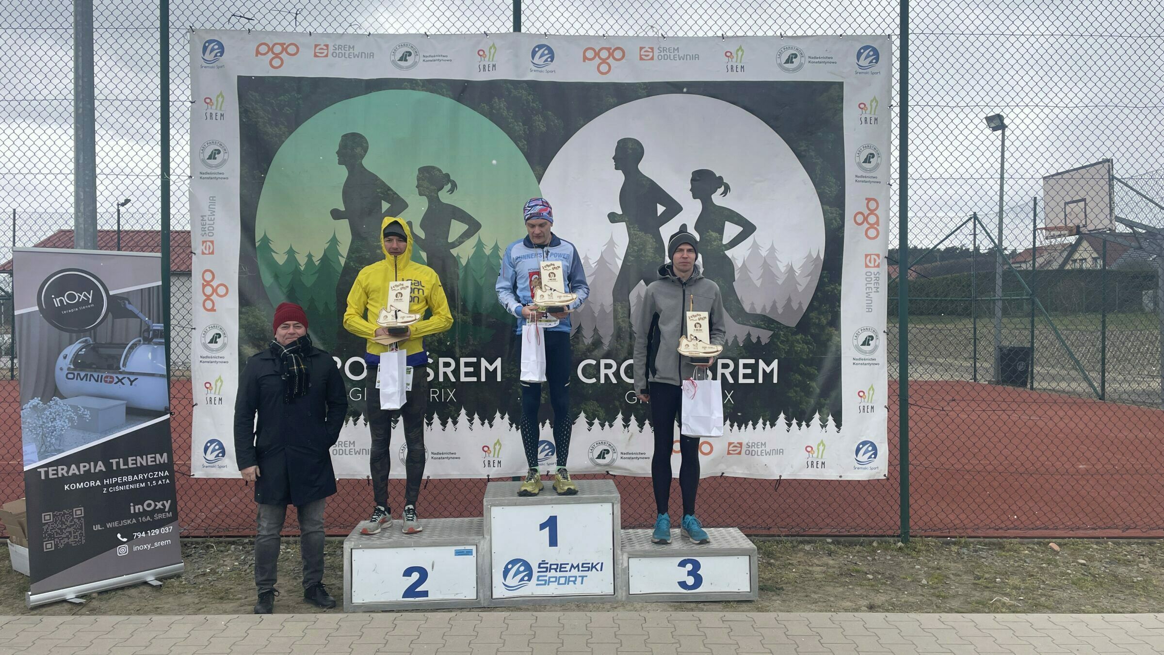 Zdjęcie przedstawia ceremonię wręczenia nagr&oacute;d po biegu w&nbsp;ramach wydarzenia Cross Śrem Grand Prix. Trzech mężczyzn stoi na&nbsp;podium z&nbsp;nagrodami w&nbsp;dłoniach, a&nbsp;za nimi rozciąga się duży baner z&nbsp;graficznym motywem biegu przełajowego &ndash; sylwetkami biegnących kobiety i&nbsp;mężczyzny na&nbsp;tle lasu. U g&oacute;ry banera znajdują się logotypy sponsor&oacute;w i&nbsp;organizator&oacute;w, m.in. &bdquo;Śremski Sport&rdquo;, &bdquo;Śrem Odlewnia&rdquo; oraz&nbsp;&bdquo;PGO&rdquo;.  Na podium:  Na najwyższym, środkowym miejscu (1) stoi mężczyzna w&nbsp;niebieskiej bluzie z&nbsp;napisem &bdquo;MINNESOTA POWER&rdquo;, czarnych legginsach w&nbsp;białe kropki i&nbsp;ż&oacute;łtych butach biegowych. Ma na&nbsp;głowie kolorową opaskę. Trzyma w&nbsp;rękach drewniane trofeum w&nbsp;kształcie sportowej sylwetki oraz&nbsp;białą torbę z&nbsp;nagrodą.  Po lewej stronie (2. miejsce) stoi zawodnik w&nbsp;ż&oacute;łtej kurtce z&nbsp;kapturem i&nbsp;czarnych legginsach. R&oacute;wnież trzyma trofeum i&nbsp;torbę.  Po prawej stronie (3. miejsce) znajduje się zawodnik w&nbsp;szarej bluzie z&nbsp;czapką i&nbsp;czarnych legginsach. Trzyma identyczne trofeum i&nbsp;torbę jak pozostali.  Obok podium, po lewej stronie zdjęcia, stoi mężczyzna w&nbsp;czarnym płaszczu i&nbsp;czerwonej czapce pan Rafał Wachowiak &ndash; organizator prezes Sp&oacute;łki Śremski Sport. Obok niego widoczny jest roll-up reklamowy firmy inOxy, oferującej tlenoterapię w&nbsp;komorze hiperbarycznej.  Tło stanowi ogrodzony kort sportowy z&nbsp;czerwonego tartanu oraz&nbsp;fragment zabudowań mieszkalnych. Niebo jest pochmurne, pogoda chłodna.