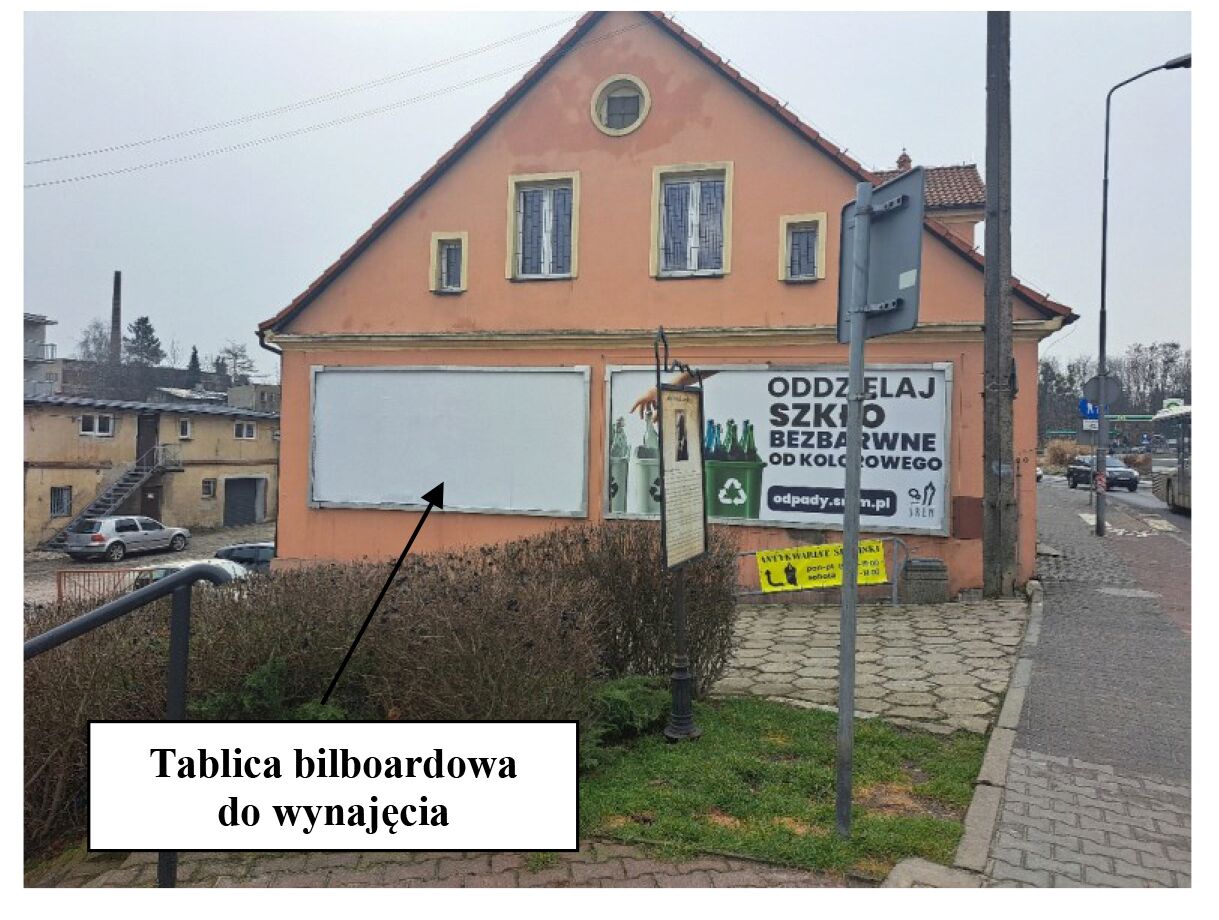 Na zdjęciu widoczna jest elewacja p&oacute;łnocna pomarańczowego budynku o&nbsp;dwuspadowym dachu, znajdującego się przy ul. Jana Kilińskiego 2 w&nbsp;Śremie. Na ścianie budynku, po lewej stronie, umieszczona jest pusta tablica bilboardowa o&nbsp;białym tle, przeznaczona do wynajęcia &ndash; wskazuje na&nbsp;nią czarna strzałka z&nbsp;podpisem &bdquo;Tablica bilboardowa do wynajęcia&rdquo;. Obok, po prawej stronie, znajduje się druga tablica z&nbsp;kolorową grafiką kampanii ekologicznej dotyczącej segregacji szkła. W tle widać inne budynki oraz&nbsp;fragment ulicy z&nbsp;chodnikiem, znakiem drogowym i&nbsp;przejeżdżającym autobusem. Pogoda jest pochmurna, a&nbsp;roślinność wok&oacute;ł budynku jest niska.